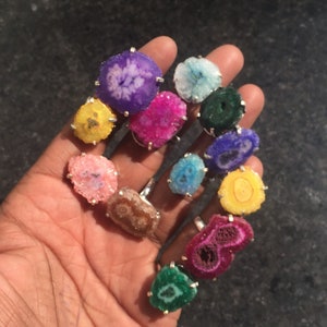 May include: A collection of 12 colorful agate rings with silver settings. The rings feature a variety of colors, including purple, pink, blue, green, and yellow. The rings are all different shapes and sizes.