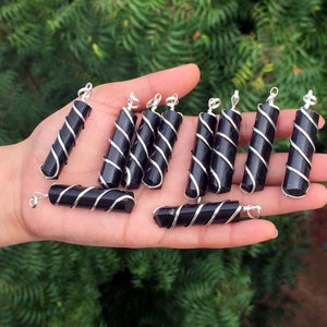 Puede incluir: Una colecci&oacute;n de colgantes de obsidiana negra envueltos en alambre de plata. Cada colgante es un cilindro largo y delgado con forma facetada. Los colgantes se muestran en una mano.