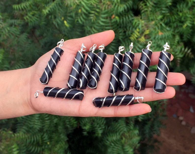 Puede incluir: Una colecci&oacute;n de colgantes de obsidiana negra envueltos en alambre de plata. Cada colgante es un cilindro largo y delgado con forma facetada. Los colgantes se muestran en una mano.
