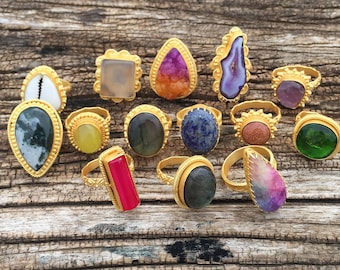 May include: A collection of gold-toned rings with various gemstones, including a shell, agate, and quartz. The rings feature different shapes and colors, such as pink, purple, green, and blue. The rings are displayed on a rustic wooden surface.