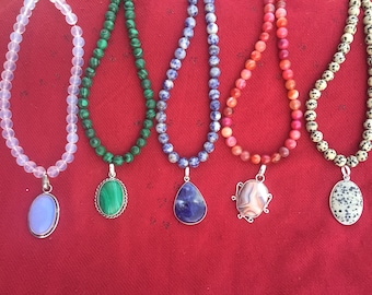 Può includere: Cinque collane con perline di pietre preziose di diversi colori e ciondoli. Le collane sono su uno sfondo rosso. I ciondoli sono in malachite, lapislazzuli, agata e diaspro dalmata.