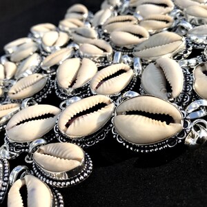Pode incluir: Um close-up de um grupo de pingentes de concha de cauri com detalhes prateados. As conchas são brancas e têm uma textura natural e orgânica. Os detalhes prateados são intrincados e detalhados, adicionando um toque de elegância aos pingentes.