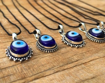 Puede incluir: Siete colgantes de plata con ojo turco azul y blanco de vidrio en cordones negros. Los colgantes están sobre una superficie de madera.