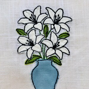 Può includere: Immagine ricamata di gigli bianchi con dettagli neri e foglie verdi in un vaso azzurro. La composizione floreale è ambientata su uno sfondo bianco, creando un design semplice ma elegante.