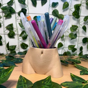 May include: A beige ceramic vase shaped like a human torso, holding a variety of colorful pens and pencils.