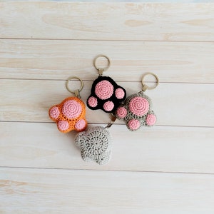 May include: Four crocheted paw print keychains in different colors: orange, black, gray, and pink. Each keychain has a silver key ring.