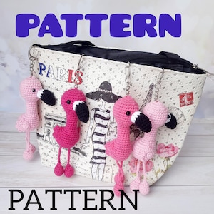Könnte beinhalten: Gehäkelte Flamingo-Schlüsselanhänger in Rosa und Fuchsia mit schwarzen Schnäbeln und roten Füßen. Die Schlüsselanhänger sind an einer Tasche mit Pariser Thema befestigt. Das Wort "PATTERN" ist in Blau und Schwarz geschrieben.