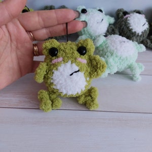 May include: A green crocheted frog with white fur on its belly and black eyes. The frog is holding a small black string.