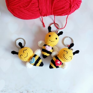 May include: Three crocheted bee keyrings with yellow bodies, black stripes, and white wings. Each bee has a heart-shaped design on its chest, with one heart being pink, one blue, and one red.