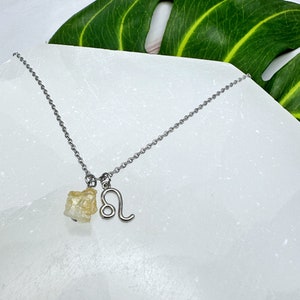 May include: A silver chain necklace with a small, clear crystal charm and a silver Leo zodiac symbol charm.