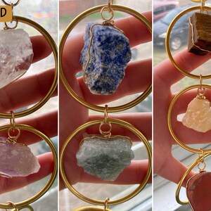 K&ouml;nnte beinhalten: Ein goldfarbener Metall-Hoop-Mobile mit sechs verschiedenen Kristallsteinen. Die Kristalle sind klar, blau, braun, rosa, gr&uuml;n und rot. Das Mobile h&auml;ngt an einer goldfarbenen Kette.