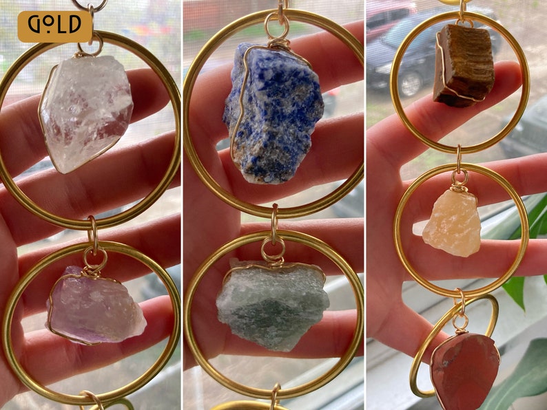 K&ouml;nnte beinhalten: Ein goldfarbener Metall-Hoop-Mobile mit sechs verschiedenen Kristallsteinen. Die Kristalle sind klar, blau, braun, rosa, gr&uuml;n und rot. Das Mobile h&auml;ngt an einer goldfarbenen Kette.