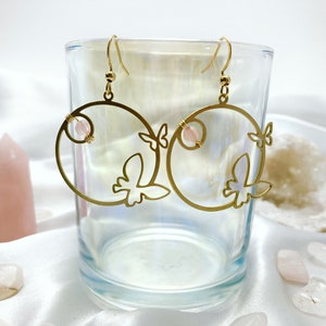 May include: Gold hoop earrings with a delicate butterfly design and a pink gemstone accent. The earrings are displayed in a clear glass container.
