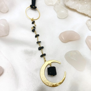 May include: A gold crescent moon charm with a black tourmaline crystal hanging from a gold chain with black tourmaline beads. The charm is surrounded by pink and white stones.