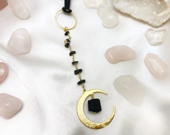 May include: A gold crescent moon charm with a black tourmaline crystal hanging from a gold chain with black tourmaline beads. The charm is surrounded by pink and white stones.