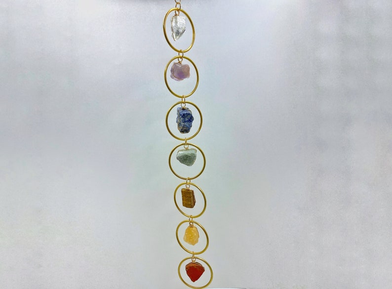 K&ouml;nnte beinhalten: Eine goldene Kette mit sieben Kristallsteinen in verschiedenen Farben, die von einer wei&szlig;en Decke h&auml;ngen. Die Steine sind klar, lila, blau, gr&uuml;n, braun, gelb und rot.