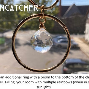 K&ouml;nnte beinhalten: Ein goldener Sonnenf&auml;nger mit einem Kristallprisma, das an einem Ring h&auml;ngt. Der Sonnenf&auml;nger ist so konzipiert, dass er Regenb&ouml;gen erzeugt, wenn Sonnenlicht durch ihn scheint. Der Text "SUNCATCHER" ist oben im Bild sichtbar.