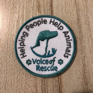 Puede incluir: Un parche bordado blanco con un borde verde azulado. El parche presenta una silueta de perro estilizada y el texto "Helping People Help Animals Voice of Rescue".