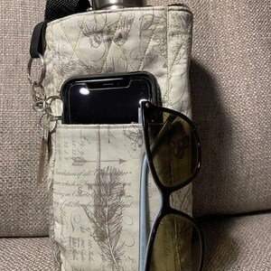 May include: A quilted fabric water bottle holder with a black strap and key ring. The holder has a pocket containing a smartphone and sunglasses. A stainless steel Klean Kanteen bottle is visible at the top. The fabric has a vintage print.