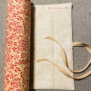 May include: A cylindrical fabric roll with a cream background and a red floral pattern, featuring beige ribbon ties at the ends. Attached is a flat, rectangular cream fabric flap with a faint script print and the embroidered name Kathy.