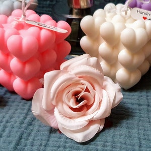 May include: Three heart-shaped cube candles in pink, white, and purple. The candles are decorated with a small tag that says "Handmade with love".