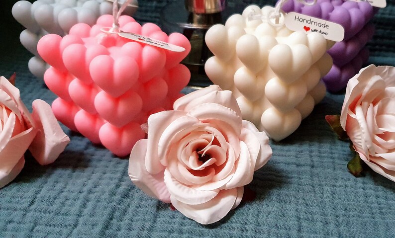 May include: Three heart-shaped cube candles in pink, white, and purple. The candles are decorated with a small tag that says "Handmade with love".