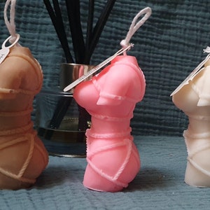 May include: Three decorative candles shaped like a female torso, each in a different color: brown, pink, and white. The candles are tied with rope and have a tag that says "Handmade".