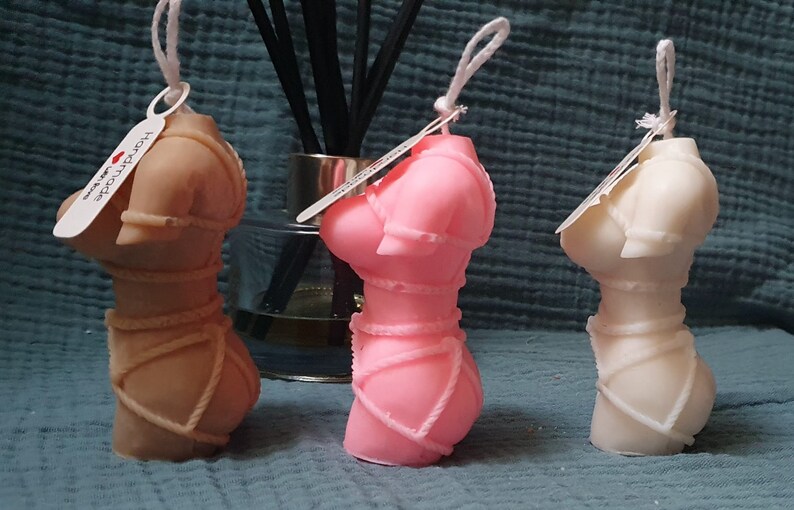May include: Three decorative candles shaped like a female torso, each in a different color: brown, pink, and white. The candles are tied with rope and have a tag that says "Handmade".