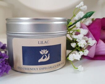 May include: A silver-colored tin candle labeled "LILAC" with a blue and beige label featuring a lotus flower design. The text "ECO FRIENDLY ESPECIALLY FOR YOU" is below the label. Purple flowers and a pink rose are next to the candle.