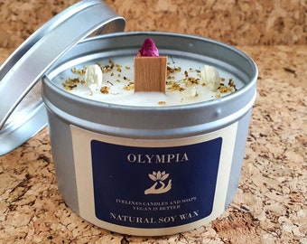 May include: A silver tin candle with a white wax top and a label that reads "OLYMPIA IVELINAS CANDLES AND SOAPS VEGAN IS BETTER NATURAL SOY WAX". The candle is decorated with dried flowers and a wooden wick.