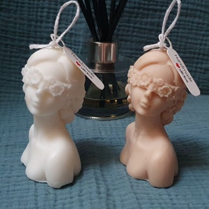May include: Two white and beige sculpted bust candles with blindfolds made of floral details. The candles are handmade and have tags that say "Handmade with Love".