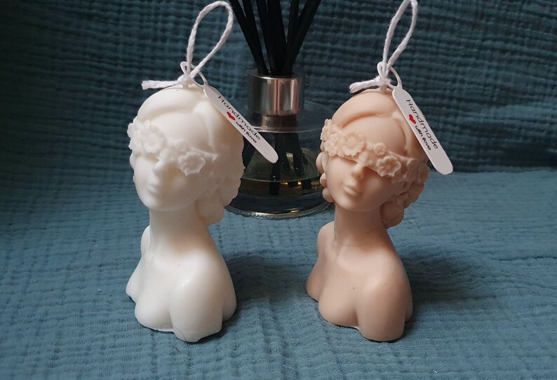 May include: Two white and beige sculpted bust candles with blindfolds made of floral details. The candles are handmade and have tags that say "Handmade with Love".