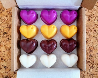 May include: A box of heart-shaped wax melts in various colors: purple, yellow, brown, and white. Each melt has a small hole at the top. The box is made of cardboard and sits on a corkboard surface.
