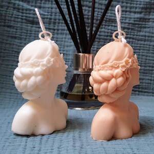 May include: Two white and beige sculpted bust candles with a braided hairstyle and a floral headband. The candles are on a blue textured surface.