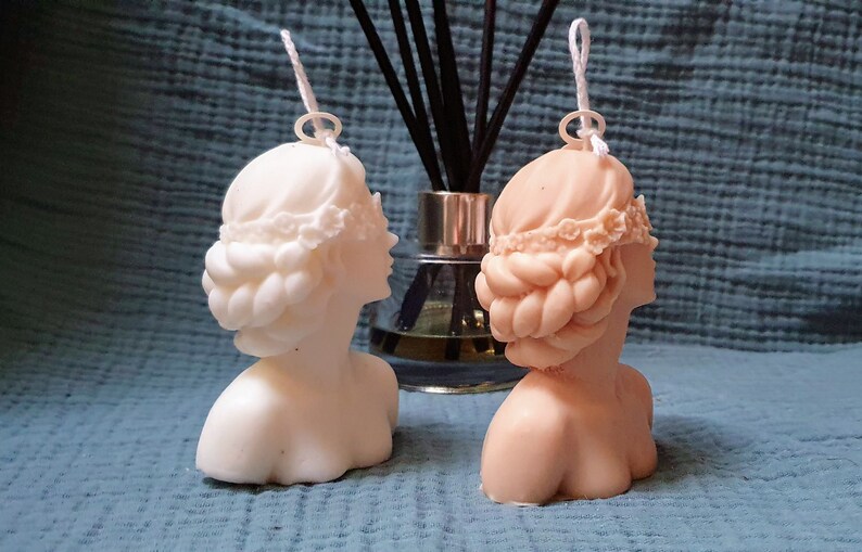 May include: Two white and beige sculpted bust candles with a braided hairstyle and a floral headband. The candles are on a blue textured surface.