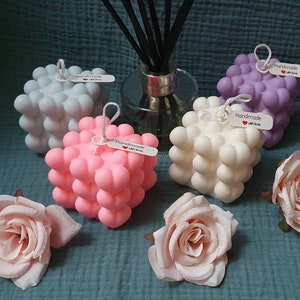 May include: Four cube-shaped candles with a textured surface, each in a different color: gray, pink, white, and purple. Each candle has a small tag that says "Handmade with love". The candles are arranged on a blue and purple textured surface with two pink roses.