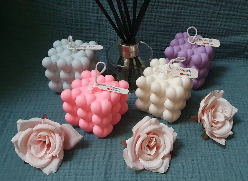 May include: Four cube-shaped candles with a textured surface, each in a different color: gray, pink, white, and purple. Each candle has a small tag that says "Handmade with love". The candles are arranged on a blue and purple textured surface with two pink roses.
