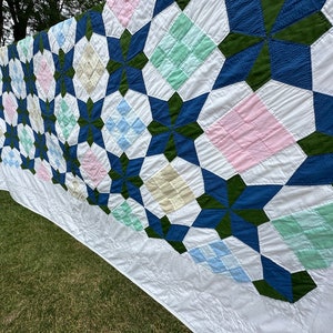 May include: A white quilt with a geometric pattern in shades of blue, green, pink, and cream. The quilt is hanging on a clothesline outdoors.