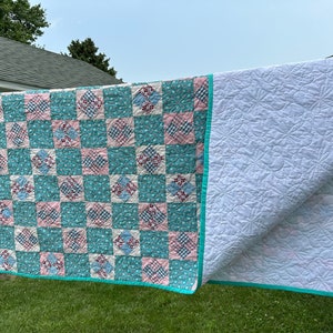 May include: A patchwork quilt with a teal, pink, and white checkered pattern. The quilt is hanging on a clothesline outside. The back of the quilt is white with a quilted pattern.