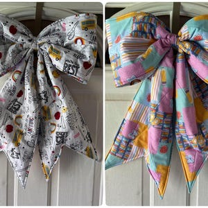 May include: Two decorative fabric bows. One bow is white with school-themed graphics and text. The other bow is a patchwork design in pink, blue, and yellow. Both bows are hanging on a white door.