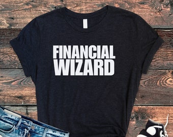 Könnte beinhalten: Ein schwarzes T-Shirt mit dem Aufdruck "FINANCIAL WIZARD" in weiß auf der Vorderseite. Das Shirt liegt auf einer Holzoberfläche mit einer blauen Jeans und einem Paar weißen Sneakers.