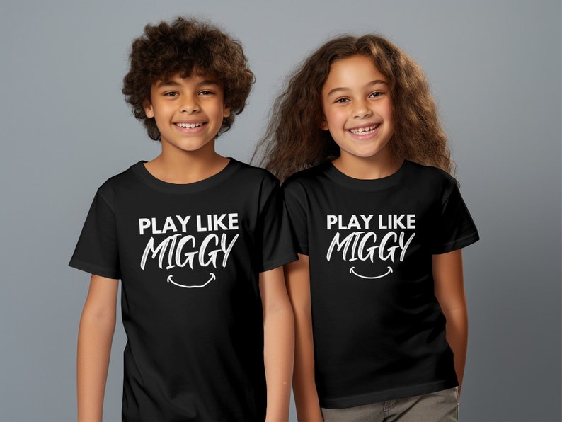 May include: Two children wearing black t-shirts with white text that says "Play Like Miggy" and a smiley face.