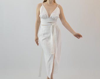 May include: A white linen two-piece outfit featuring a V-neck crop top and a high-waisted wrap skirt with a side slit. The outfit includes a matching belt. The model is wearing black sandals.