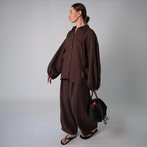 Può includere: Un completo in lino marrone composto da una camicia abbottonata a maniche lunghe con dettagli a nappe e pantaloni a gamba larga. L'outfit è abbinato a sandali neri e una borsa nera intrecciata.