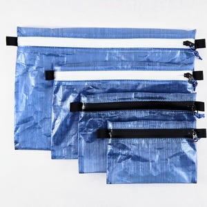 Op de afbeelding: Een set van vier rechthoekige blauwe etuis met een gekreukte textuur. De twee grotere etuis hebben witte ritssluitingen, terwijl de twee kleinere etuis zwarte ritssluitingen hebben. Elk etui is voorzien van zwarte lipjes aan de zijkanten.
