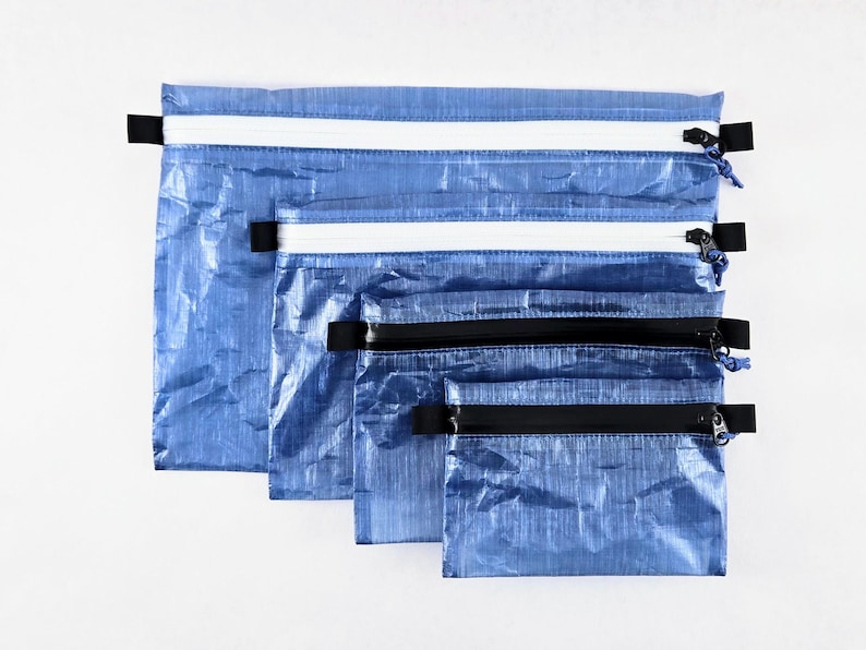 Op de afbeelding: Een set van vier rechthoekige blauwe etuis met een gekreukte textuur. De twee grotere etuis hebben witte ritssluitingen, terwijl de twee kleinere etuis zwarte ritssluitingen hebben. Elk etui is voorzien van zwarte lipjes aan de zijkanten.