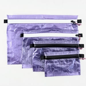Op de afbeelding: Een set van vier rechthoekige etuis in verschillende maten, gemaakt van een gekreukt, doorschijnend paars materiaal. Elk etui is voorzien van een ritssluiting met een treklipje, waarbij de twee grotere etuis witte ritsen hebben en de twee kleinere zwarte.