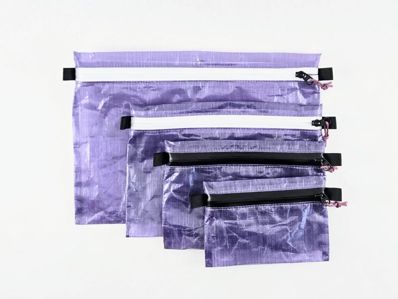 Op de afbeelding: Een set van vier rechthoekige etuis in verschillende maten, gemaakt van een gekreukt, doorschijnend paars materiaal. Elk etui is voorzien van een ritssluiting met een treklipje, waarbij de twee grotere etuis witte ritsen hebben en de twee kleinere zwarte.