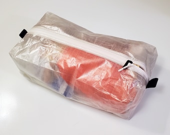 May include: A clear, translucent zippered pouch with a white zipper and black pull tabs. The pouch is filled with various items, including a red, translucent object.