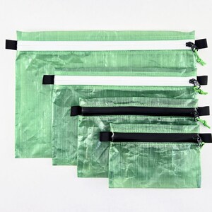 Op de afbeelding: Een set van vier rechthoekige etuis in verschillende maten, gemaakt van een gekreukt, doorschijnend lichtgroen materiaal. Elk etui is voorzien van een horizontale ritssluiting met een zwarte trekker en een klein groen koordje.